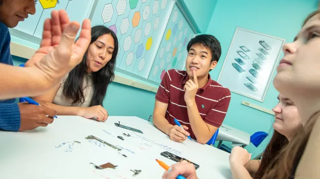 Five teenagers sitting around a table that is covered with paper. Some of the teens are holding magic markers. A disassembled cellphone is on the table and there is writing on the paper near the cellphone parts.