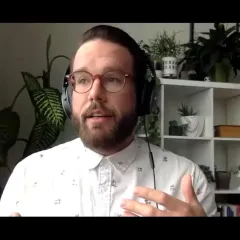Climate scientist Jeremy Hoffman, a light-skinned, bearded man wearing headphones and white shirt, sitting in front of a bookcase with plants on it