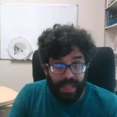 A man sitting in a chair. He has black, curly hair and a black beard and mustache, and he is wearing glasses.