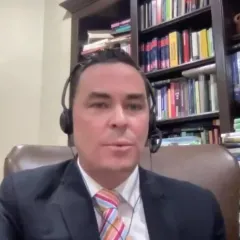 A man in a dark jacket, white shirt, and colorful striped tie sitting in a chair with a full bookcase behind him. He has a tan complexion, short dark hair, and is wearing a headphones with a headset microphone.