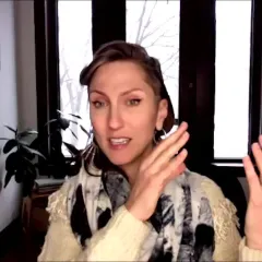 A woman sitting in front of a window with a plant. She has shoulder-length brown hair and is wearing a black and white scarf over a cream-colored sweater. She is gesturing with both hands as she speaks.