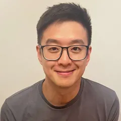 A smiling man with dark hair and glasses with medium skin tone in front of an off-white backdrop