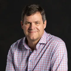 A headshot of Kirk Johnson, a white man with short brown hair wearing a blue and red gingham button-up shirt.