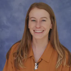 Light-skinned woman with long hair in a collared orange shirt in front of a blue background