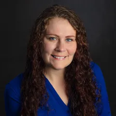 A woman in a blue dress smiling for a posed headshot photograph against a dark gray backdrop