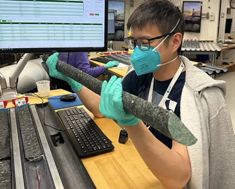 A man wearing a turquoise face mask and gloves holds a long, half-cylinder of rock next to a computer monitor and in front of several trays of similar rocks.