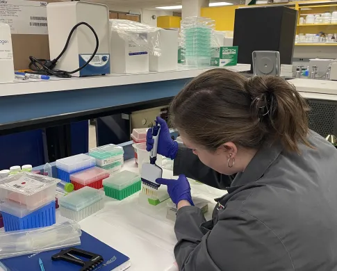 A woman in a grey lab coat sits at a bench surrounded by scientific implements. She holds an 8-tipped scientific pipette and is transferring liquid to a translucent gridded plate.