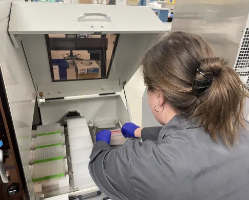 A woman wearing a grey lab coat and purple latex gloves arranges rows of plates and trays containing liquid on a stainless steel platform below an open hatch with a handle
