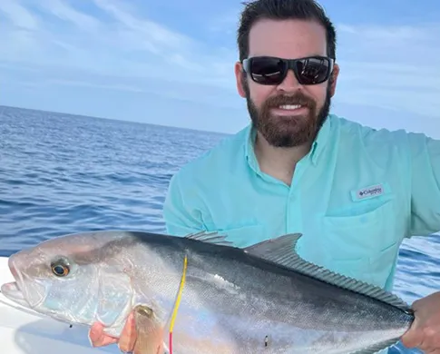 A man wearing sunglasses and a turquoise shirt and holding a large fish in front of him, with the ocean behind him.