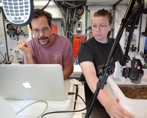 A man and woman in an industrial looking room look at a computer screen sitting next to a camera tripod. The woman guides a tray of water containing a colorful square plate.