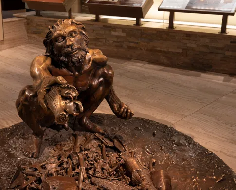 Bronze sculpture of an early human with a beard and mustache squatting and holding out a piece of wood. 