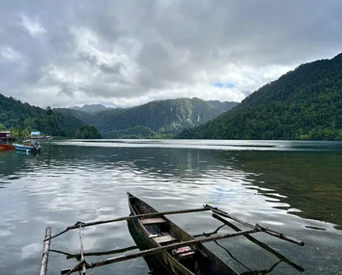 A double-outrigger canoe in a body of water with piers on the left shore and tree-covered mountains in the distance. The sky is full of dark clouds.
