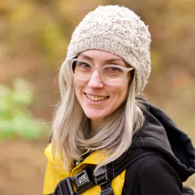 A woman smiling for a photograph wearing a beanie, glasses, and a jacket