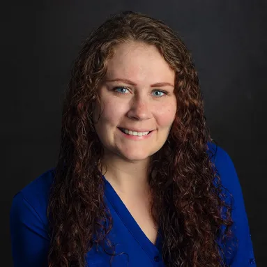 A woman in a blue dress smiling for a posed headshot photograph against a dark gray backdrop