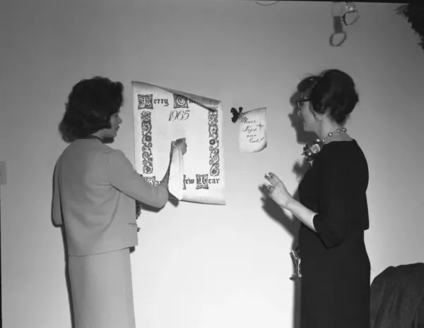 Black and white photo of two women signing an attendance sheet for a holiday party dated 1965