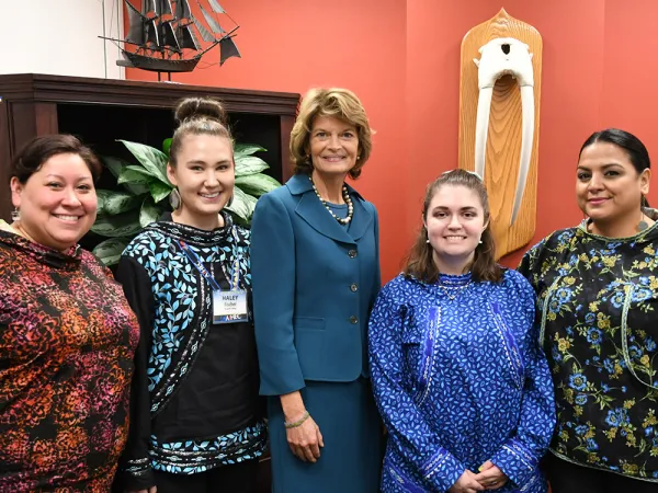 Five women standing in front of a bookcase and a salmon-colored wall with walrus tusks displayed on it. The woman in the middle is U.S. Senator Lisa Murkowski.