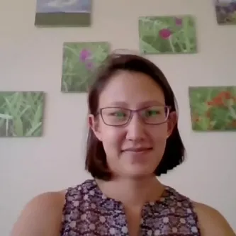 A medium-light-skinned woman with dark hair and glasses, wearing a patterned sleeveless top, sitting in front of a wall with seven small, unframed paintings on it