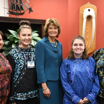 Five women standing in front of a bookcase and a salmon-colored wall with walrus tusks displayed on it. The woman in the middle is U.S. Senator Lisa Murkowski.