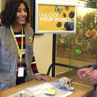 A teen girl in a gray vest standing at a cart with collection objects on it. She is talking to two visitors.