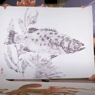 A man and a woman standing behind a table and holding up a large black and white print of a fish between them