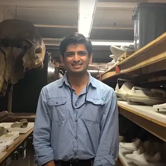 A medium-skinned man with black hair, wearing a blue button-down shirt standing between some shelves and cabinets with two large, elephant-like fossil skulls behind him and to his left.