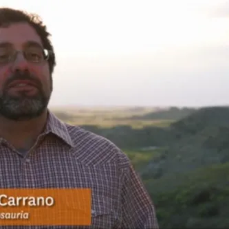 Matt Carrano, a man with dark hair,  beard and mustache, standing in front of a landscape of rocky hills and grasslands.