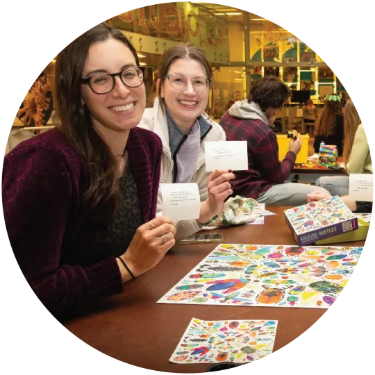 Two women sitting at a table with a completed jigsaw puzzle of colorful beetles in front of them. The woman are holding up white cards with typewritten messages on them.