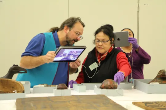 Man holding an iPad using video to show objects; headphones are connected to a woman explaining the objects.