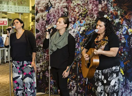 Three women standing at microphones singing; individual on the right has a guitar.