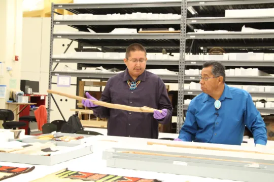 One man holding a traditional Zuni digging stick while another looks on.