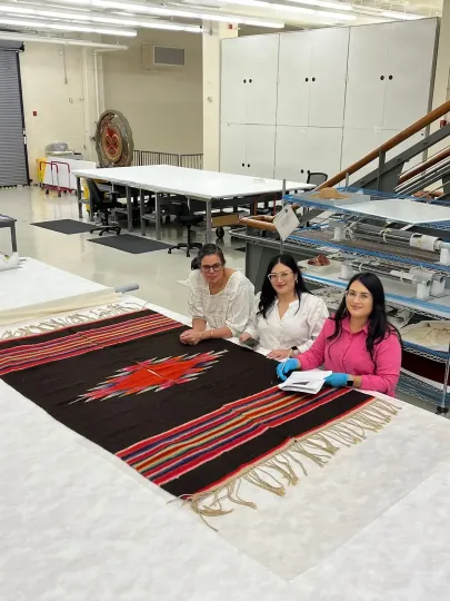 Three women standing behind a large viewing table that has a large black poncho, decorated with a red geometric pattern in the middle, alternating red, blue, white, green, and yellow stripes on both ends, and tan fringe at the edge of the poncho.