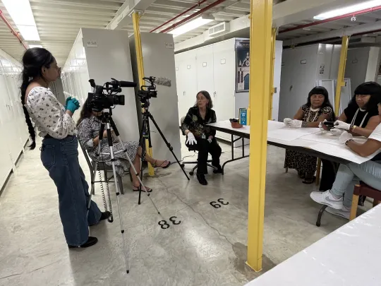 A group of Wauja women sitting around a table while two other women are video and audio recording their conversation.