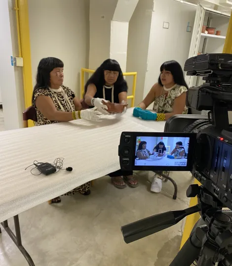 Three women sitting at a table examining a ceramic pot; same image seen through a camera in the foreground.