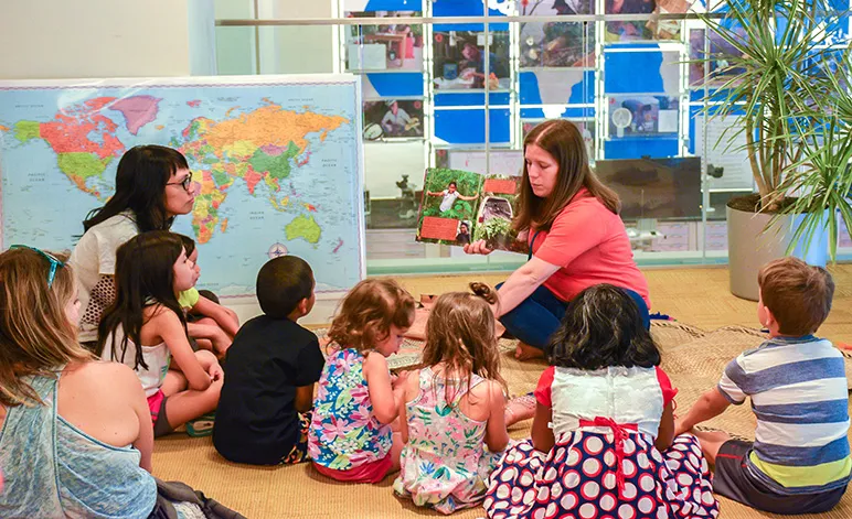 A woman sitting on the floor next to a word map reads a book to six children sitting in front of her.