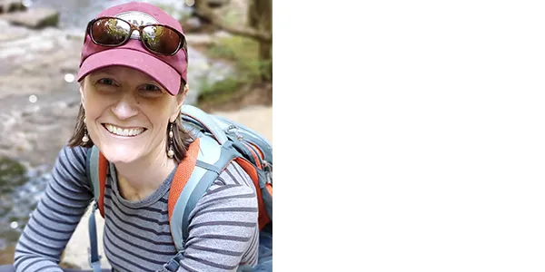 Portrait of Amy Freestone, a light-skinned woman wearing sunglasses on a cranberry-colored baseball cap, as well as a striped blue shirt and a backpack.