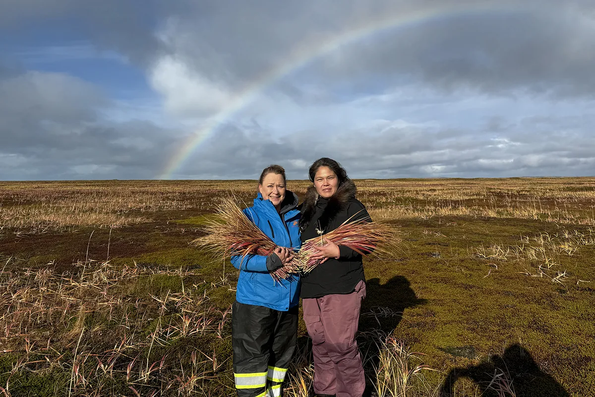 Two women in winter coats standing in a field of reddish-brown grass with a rainbow behind them. They are holding bundles of the reddish-brown grass.