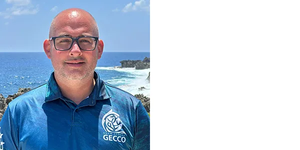 A smiling medium-light-skinned man with very short gray-and-dark hair and a gray-and-dark short mustache and beard. He is wearing dark-framed glasses and a collared, blue shirt with the words Guam Epscon Gecco on the left chest. Behind and below him is the ocean and a coastline.