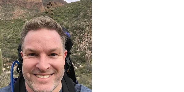 A smiling light-skinned man with short dirty-blond hair and a short gray-and-dark mustache and beard, standing in a hilly, rocky landscape with two cacti. He is wearing a blue shirt and a backpack. 