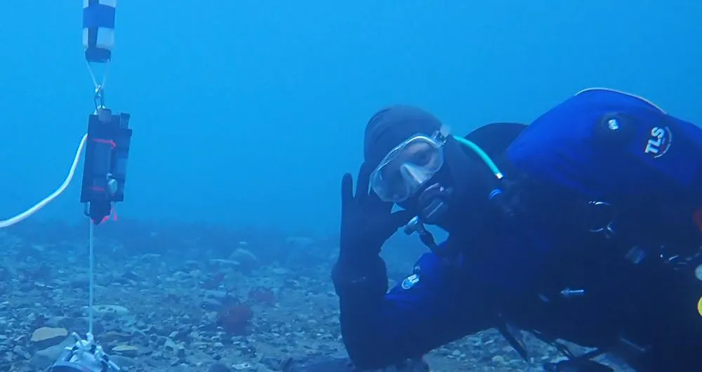 A scuba diver rests his elbow and leg on the sea floor next to a data logger tethered to a weight on the bottom.