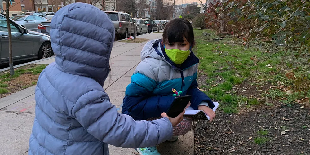 Two children standing next to a lawn by a sidewalk. One child is showing their smart phone to the other.