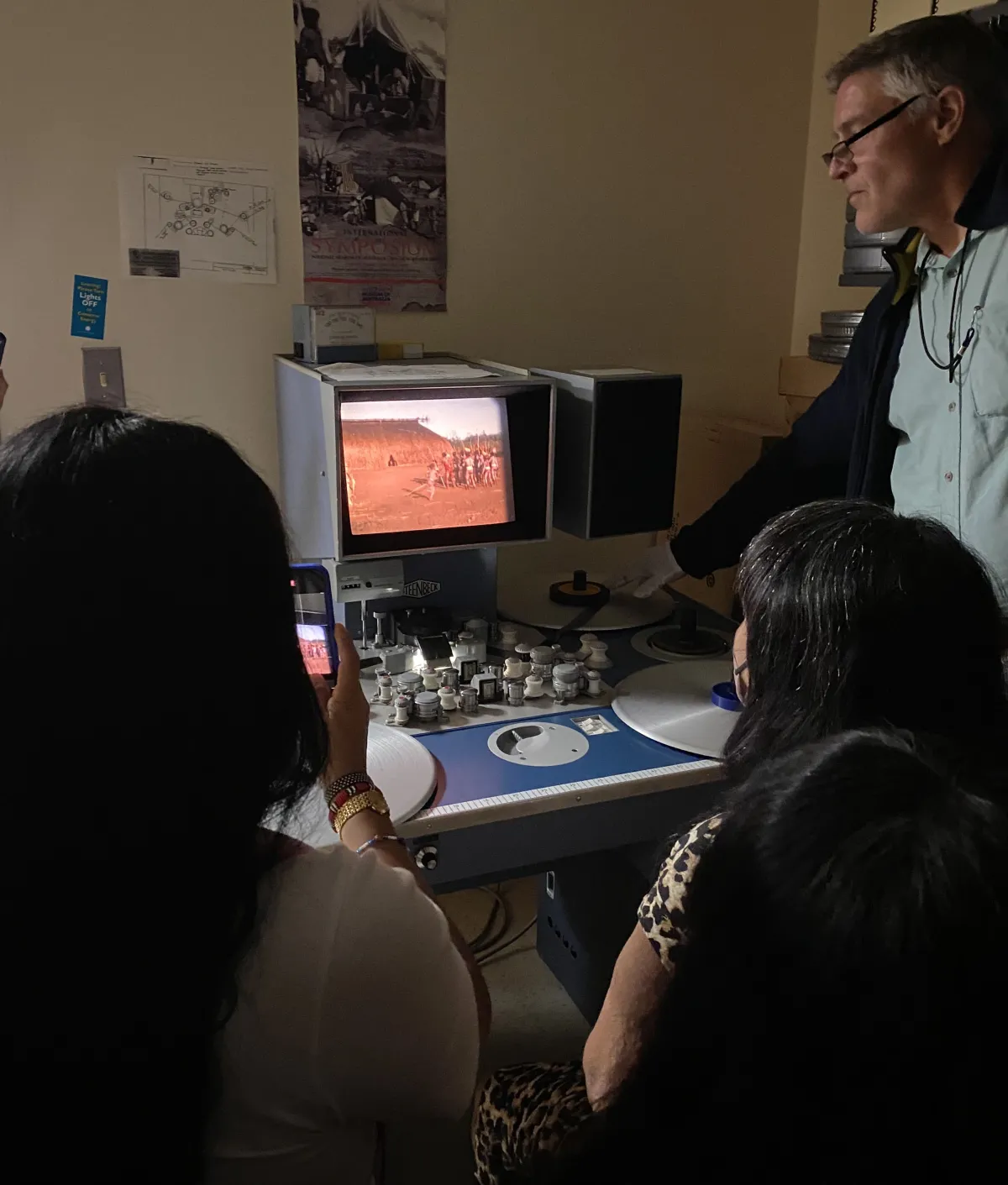 Two women looking at a screen with one taking a picture; a man is in the background controlling the viewing screen.
