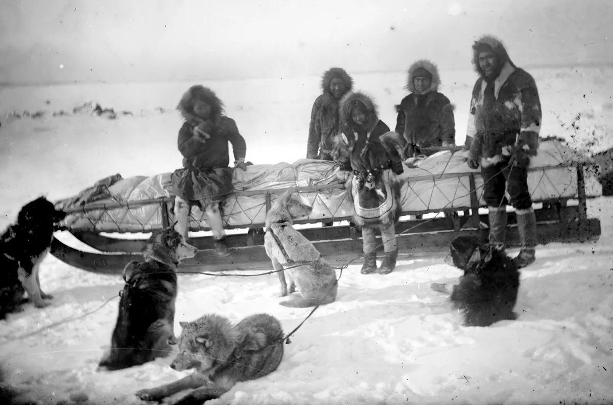 Black and white image of five Iñupiaq people wearing parkas and gathered around and on a large sled in a snow-covered landscape. There are five sled dogs sitting in the foreground.