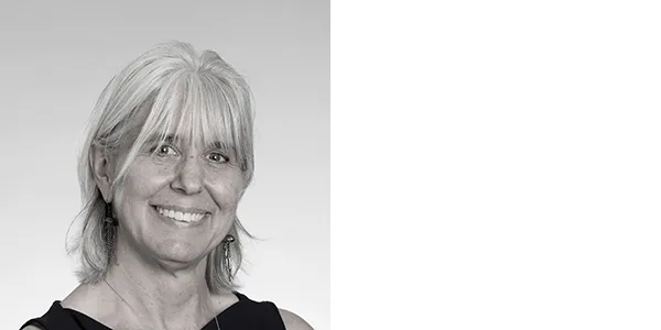 A black and white portrait of Jennifer Collins, a woman with shoulder-length white hair, who is wearing dangling earrings, a necklace, and a black sleeveless top.
