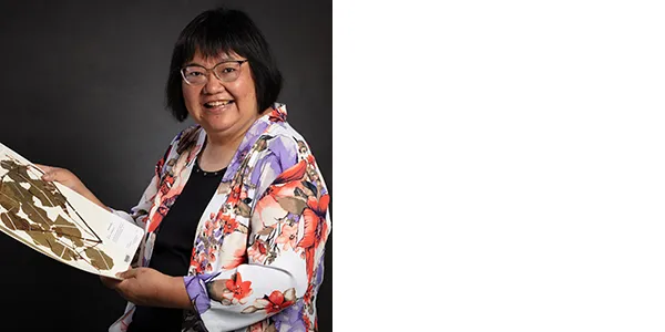 Portrait of Jun Wen, a medium-light skinned woman with short black hair, wearing glasses and a flower-print jacket over a black t-shirt. She is holding some paper, on which is a green and brown pressed plant specimen.