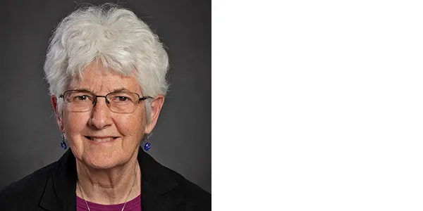 Portrait of Kay Behrensmeyer, a light-skinned woman with short white hair, who is wearing glasses, a black jacket over a magenta shirt, a necklace with a silver pendant, and blue earrings.
