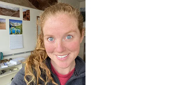 A smiling, light-skinned woman with long blonde hair and blue-gray eyes. She is wearing a gray fleece over a red shirt. Behind her is a calendar and some photos of desert landscapes.