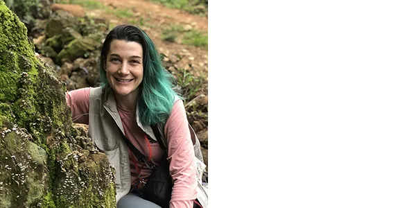 A smiling, medium-light-skinned woman with black and emerald green hair, wearing a thin gray vest over a long-sleeved pink t-shirt. She is sitting next to a large rock covered with green, brown, and white matter which may be lichens or fungi.