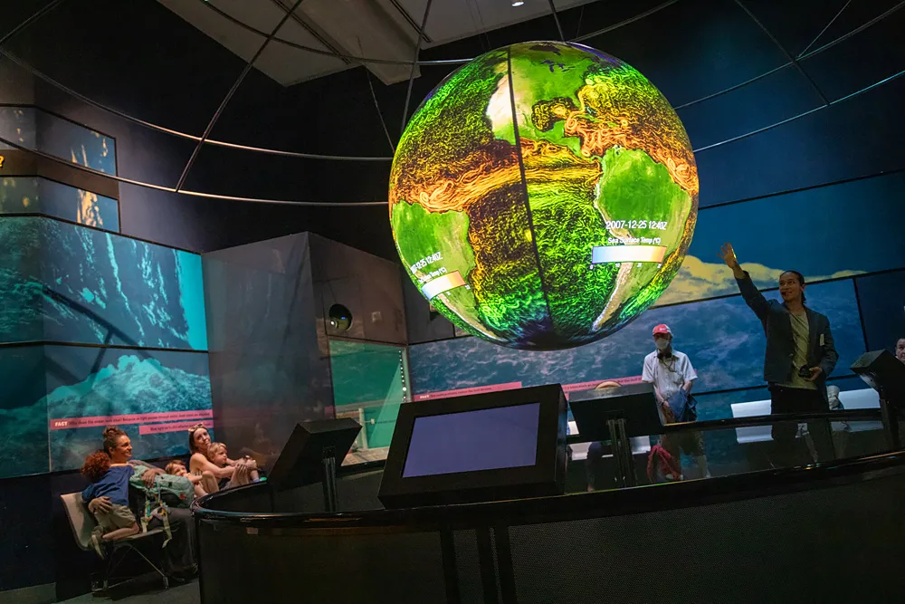 Eight people in an exhibit hall looking up at a large globe suspended from the ceiling. The globe displays a map of the world in green, brown, yellow, and white. A man stands near the globe, pointing at it while talking.