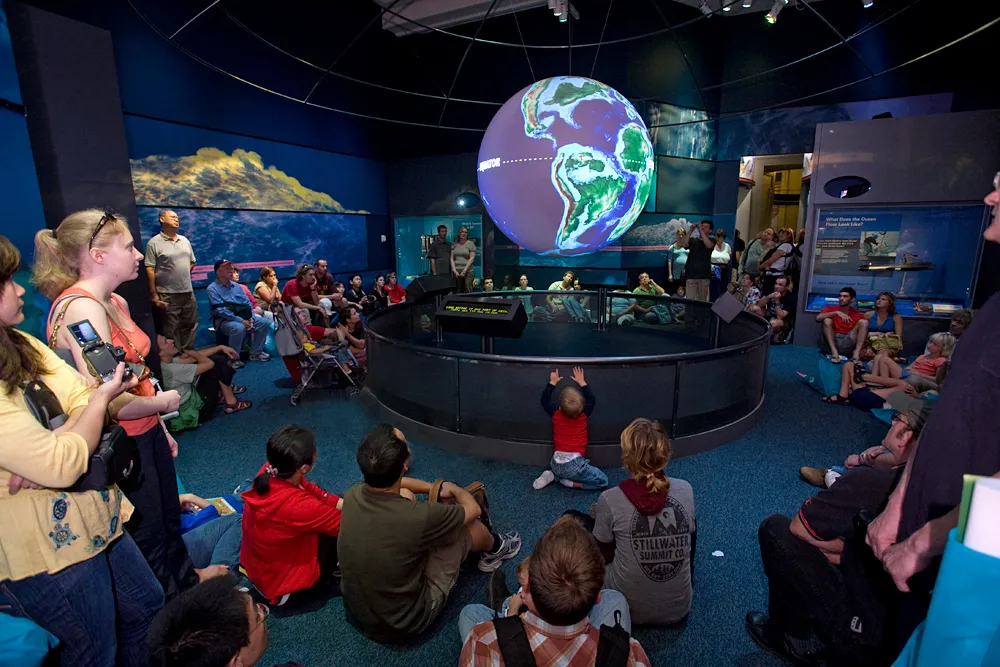 A room with a large, illuminated globe of the Earth hanging from the ceiling, with dozens of people sitting on floor and standing around it.