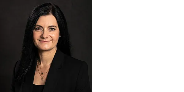 A smiling, medium-light-skinned woman with long black hair and brown eyes. She is wearing a black jacket over a black top, and two necklaces, one with a pendant of the letter R.